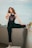 A woman with long, wavy hair is posing confidently on an urban rooftop. She is wearing a sleeveless top with the phrase 'Live Life Loud' and black sweatpants. One of her legs is raised and resting on a ledge, showing off her sneakers. The background features a cityscape under a soft, cloudy sky.