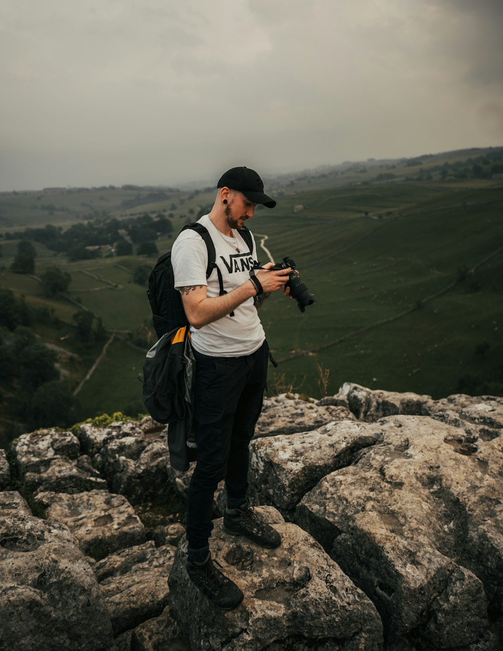 man in white t-shirt and black pants holding black dslr camera standing on rocky mountain