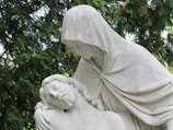 A white marble statue depicting a serene and solemn scene where one figure is cradling another. The clothed figure, with a hood over their head, gazes down compassionately at the reclining figure with closed eyes. This is set against a backdrop of lush green foliage.