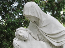 A white marble statue depicting a serene and solemn scene where one figure is cradling another. The clothed figure, with a hood over their head, gazes down compassionately at the reclining figure with closed eyes. This is set against a backdrop of lush green foliage.