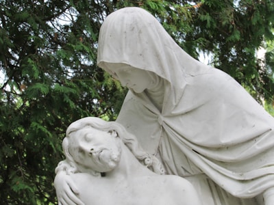 A white marble statue depicting a serene and solemn scene where one figure is cradling another. The clothed figure, with a hood over their head, gazes down compassionately at the reclining figure with closed eyes. This is set against a backdrop of lush green foliage.