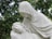 A white marble statue depicting a serene and solemn scene where one figure is cradling another. The clothed figure, with a hood over their head, gazes down compassionately at the reclining figure with closed eyes. This is set against a backdrop of lush green foliage.