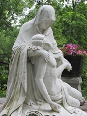 A contemplative statue depicts a hooded figure gently cradling a bare-chested, reclining individual, set against a lush, green wooded background. Vibrant pink and red flowers complement the serene and solemn scene.