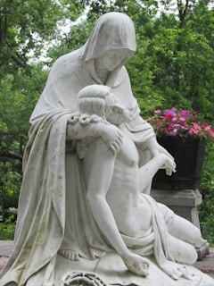 A contemplative statue depicts a hooded figure gently cradling a bare-chested, reclining individual, set against a lush, green wooded background. Vibrant pink and red flowers complement the serene and solemn scene.
