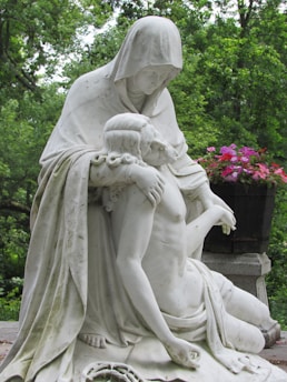 A contemplative statue depicts a hooded figure gently cradling a bare-chested, reclining individual, set against a lush, green wooded background. Vibrant pink and red flowers complement the serene and solemn scene.