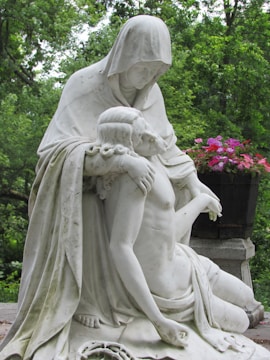 A contemplative statue depicts a hooded figure gently cradling a bare-chested, reclining individual, set against a lush, green wooded background. Vibrant pink and red flowers complement the serene and solemn scene.