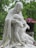 A contemplative statue depicts a hooded figure gently cradling a bare-chested, reclining individual, set against a lush, green wooded background. Vibrant pink and red flowers complement the serene and solemn scene.