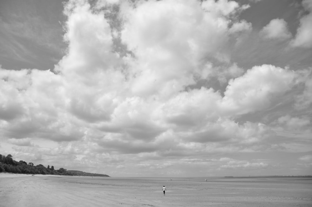grayscale photo of person walking on beach under cloudy sky