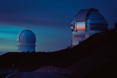 Two large observatories are positioned on a hillside under a dusky sky. The structures have domed roofs, and one of them reflects a warm glow, likely from a setting sun. The foreground is a road leading towards the observatories.