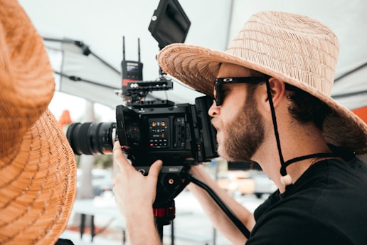 A person wearing a wide-brimmed straw hat and sunglasses is operating a professional video camera. The setting appears to be outdoors with some background blur indicating a busy environment.