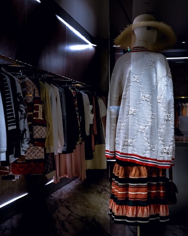 A mannequin stands in a dimly lit, upscale clothing store, wearing a white textured sweater with red and black striped details, paired with a layered skirt featuring earth tones. It dons a wide-brimmed straw hat. Clothes are neatly arranged on a rack alongside the mannequin, displaying a variety of patterns and colors.