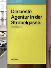 A yellow sign with bold black text in German promoting an agency located on a street. The background shows part of an urban setting with tables and chairs on a sidewalk.