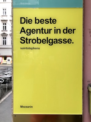 A yellow sign with bold black text in German promoting an agency located on a street. The background shows part of an urban setting with tables and chairs on a sidewalk.