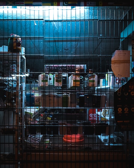 A dimly lit storefront or display cabinet is viewed through a metal grid or bars. Various electronic items or gadgets are arranged on shelves, some of which are illuminated by a faint blue light. A piece of paper with text is taped to the grid on the right side.