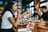A group of people sitting around a table in a restaurant, enjoying a meal together. Several dishes are spread out on the wooden table, including salads and cookies topped with colorful sprinkles. Wine glasses and a milkshake glass add to the lively dining atmosphere.