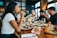 A group of people sitting around a table in a restaurant, enjoying a meal together. Several dishes are spread out on the wooden table, including salads and cookies topped with colorful sprinkles. Wine glasses and a milkshake glass add to the lively dining atmosphere.