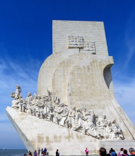 A large stone monument featuring detailed sculptures of historical figures, all facing forward while standing on the sloped base. The monument has a nautical theme, and the figures appear to represent explorers or prominent individuals. The background is a vibrant blue sky, and there are several people at the base, taking photos and observing.
