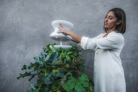 A woman wearing a white, long-sleeved top is tending to a vertical garden filled with green leafy vegetables. She is carefully adjusting a modern, white device that seems to be part of the garden setup. The background is a plain, textured gray wall which emphasizes the greenery.