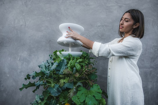 A woman wearing a white, long-sleeved top is tending to a vertical garden filled with green leafy vegetables. She is carefully adjusting a modern, white device that seems to be part of the garden setup. The background is a plain, textured gray wall which emphasizes the greenery.