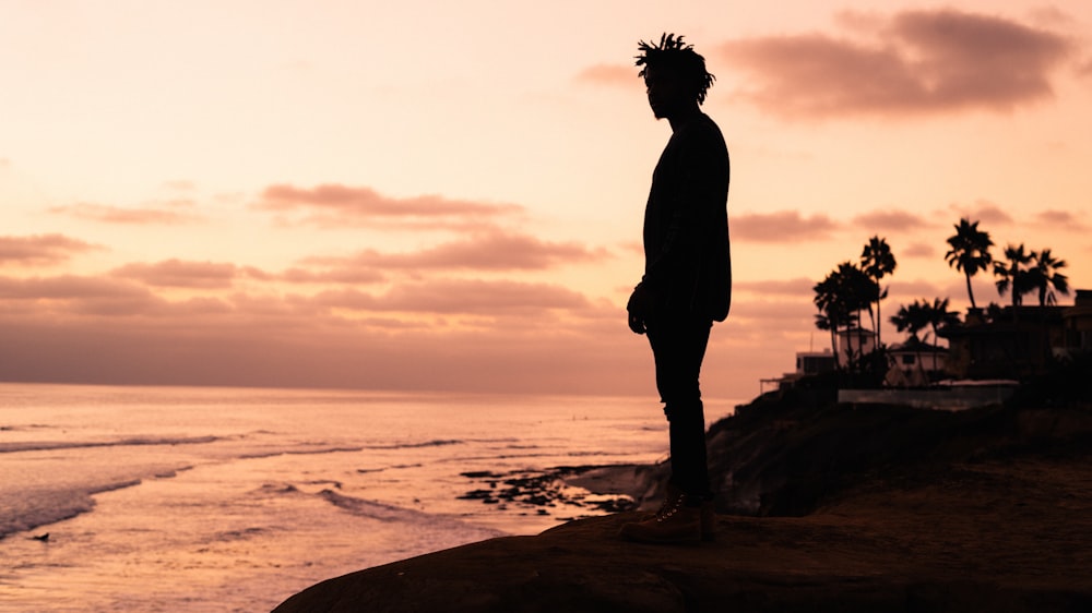 silhouette of man standing on rock formation near body of water during sunset