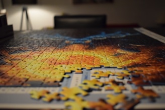A partially completed jigsaw puzzle lies on a table, with some pieces scattered around the edges. The puzzle appears to feature vibrant colors and abstract patterns. In the background, there is dim lighting and out-of-focus furniture.