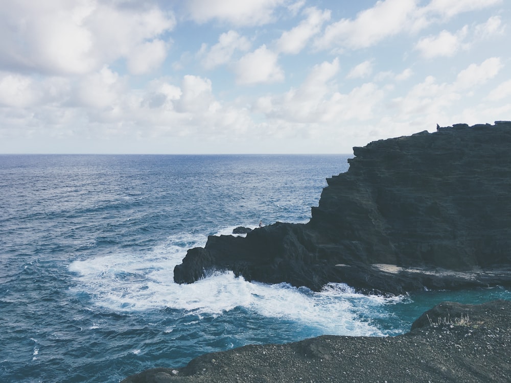 ocean waves crashing on black rock formation under white clouds and blue sky during daytime