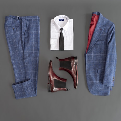 A flat lay composition featuring a stylish men's outfit. The ensemble includes a blue plaid suit jacket and matching trousers, a white dress shirt with a black tie, and a pair of brown leather Chelsea boots. The interior lining of the jacket is red, adding a touch of elegance.