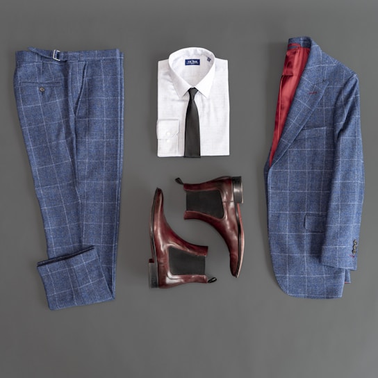 A flat lay composition featuring a stylish men's outfit. The ensemble includes a blue plaid suit jacket and matching trousers, a white dress shirt with a black tie, and a pair of brown leather Chelsea boots. The interior lining of the jacket is red, adding a touch of elegance.