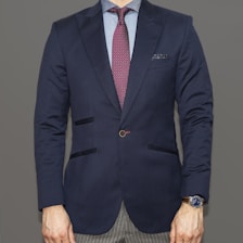 A person dressed in a formal navy blue blazer paired with a light blue shirt and a burgundy patterned tie. The bottom portion of gray pinstriped pants is visible. A watch with a blue face and metallic strap is worn on the left wrist. The attire conveys a professional and polished appearance.