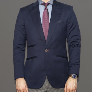 A person dressed in a formal navy blue blazer paired with a light blue shirt and a burgundy patterned tie. The bottom portion of gray pinstriped pants is visible. A watch with a blue face and metallic strap is worn on the left wrist. The attire conveys a professional and polished appearance.