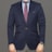A person dressed in a formal navy blue blazer paired with a light blue shirt and a burgundy patterned tie. The bottom portion of gray pinstriped pants is visible. A watch with a blue face and metallic strap is worn on the left wrist. The attire conveys a professional and polished appearance.