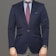 A person dressed in a formal navy blue blazer paired with a light blue shirt and a burgundy patterned tie. The bottom portion of gray pinstriped pants is visible. A watch with a blue face and metallic strap is worn on the left wrist. The attire conveys a professional and polished appearance.