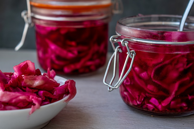 Two glass jars filled with vibrant red sauerkraut and a bowl of the same cabbage are placed on a gray surface. The jars feature metal latch lids, and a spoon is sticking out of the closer jar.