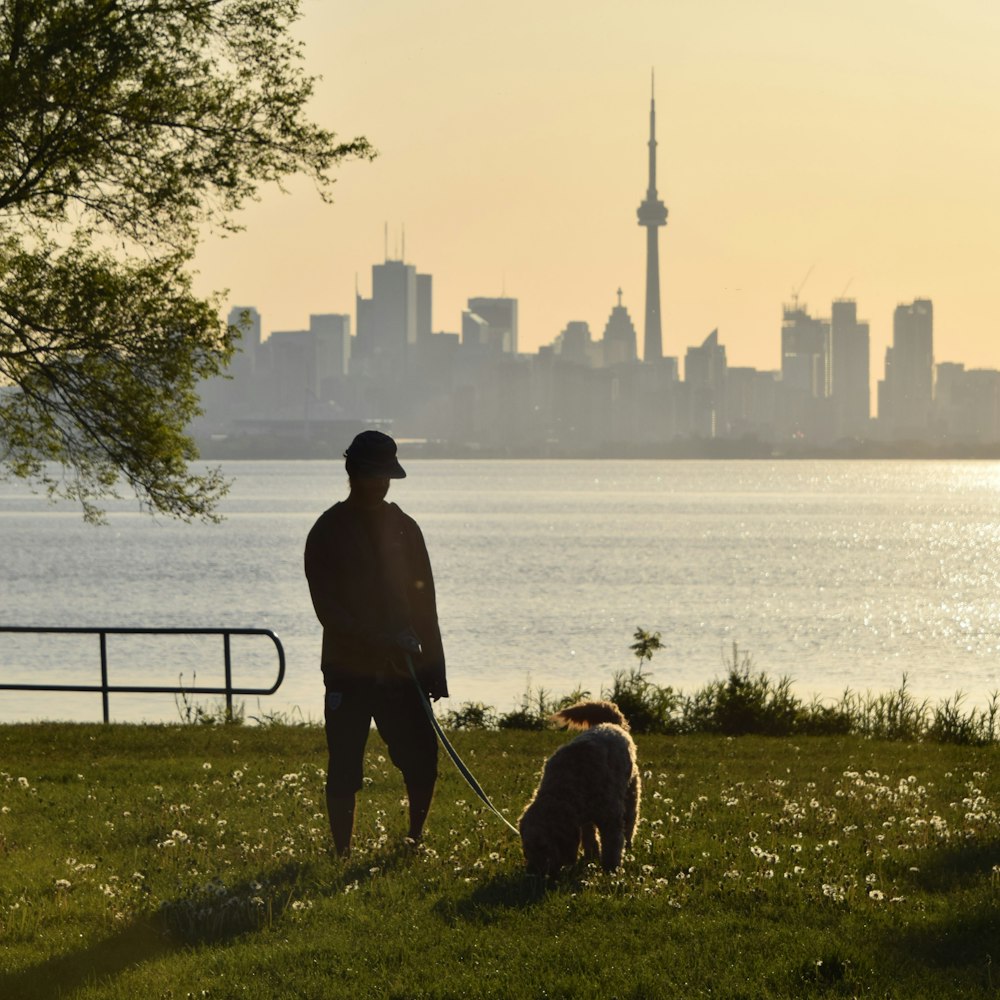 man in black jacket standing beside dog near body of water during daytime