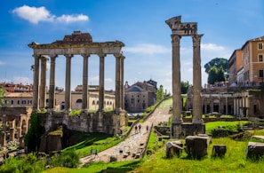 Ancient Roman ruins with tall stone columns under a bright blue sky. The ruins are surrounded by green grass and paths, with modern buildings visible in the background. Tourists walk around, exploring the historic site.