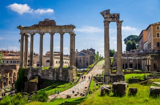 Ancient Roman ruins with tall stone columns under a bright blue sky. The ruins are surrounded by green grass and paths, with modern buildings visible in the background. Tourists walk around, exploring the historic site.