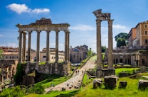 Ancient Roman ruins with tall stone columns under a bright blue sky. The ruins are surrounded by green grass and paths, with modern buildings visible in the background. Tourists walk around, exploring the historic site.