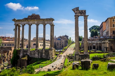 Ancient Roman ruins with tall stone columns under a bright blue sky. The ruins are surrounded by green grass and paths, with modern buildings visible in the background. Tourists walk around, exploring the historic site.