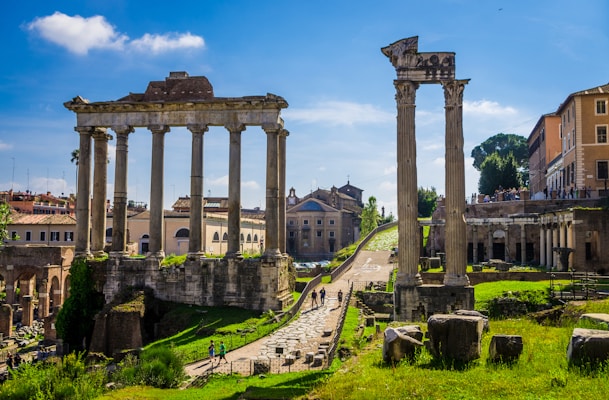 Ancient Roman ruins with tall stone columns under a bright blue sky. The ruins are surrounded by green grass and paths, with modern buildings visible in the background. Tourists walk around, exploring the historic site.