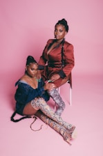 Two women pose confidently in a fashion-forward setting with a pink background. One is seated on a white stool wearing a stylish brown outfit with a deep neckline and black harness details, while the other kneels beside her in an off-the-shoulder denim top and snake-patterned thigh-high boots. Both women have intricate hairstyles and distinctive jewelry.