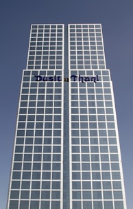 A tall, modern skyscraper with a grid of windows, featuring a sign with the name 'Dusit Thani' prominently displayed. The building has a symmetrical design with a clear blue sky in the background.