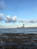 A large mosque with a tall minaret stands by the sea, under a vast blue sky with scattered clouds. In the foreground, rocky tide flats are partially submerged by water, and the cityscape stretches along the horizon.