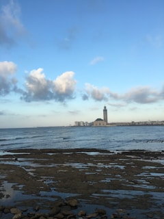 A large mosque with a tall minaret stands by the sea, under a vast blue sky with scattered clouds. In the foreground, rocky tide flats are partially submerged by water, and the cityscape stretches along the horizon.