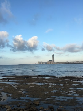 A large mosque with a tall minaret stands by the sea, under a vast blue sky with scattered clouds. In the foreground, rocky tide flats are partially submerged by water, and the cityscape stretches along the horizon.
