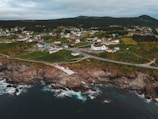 A coastal town features numerous houses dispersed across lush green fields. The landscape is bordered by rugged cliffs meeting the ocean, with waves gently lapping at the rocky shoreline. The community is connected by winding roads, and a prominent white building stands out among the houses.