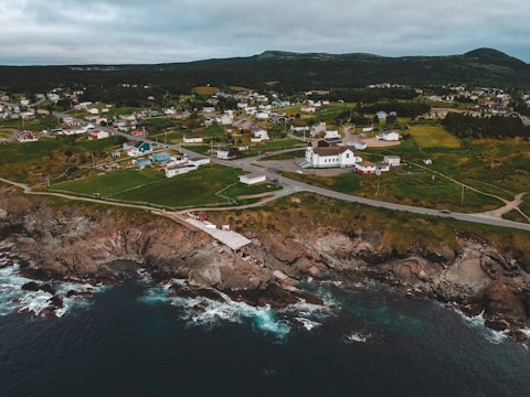 A coastal town features numerous houses dispersed across lush green fields. The landscape is bordered by rugged cliffs meeting the ocean, with waves gently lapping at the rocky shoreline. The community is connected by winding roads, and a prominent white building stands out among the houses.