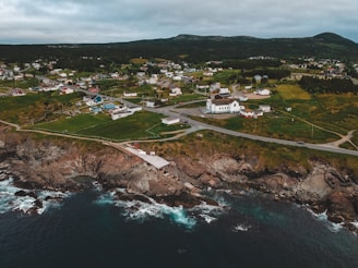 A coastal town features numerous houses dispersed across lush green fields. The landscape is bordered by rugged cliffs meeting the ocean, with waves gently lapping at the rocky shoreline. The community is connected by winding roads, and a prominent white building stands out among the houses.