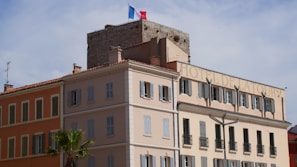 A multi-story building with a light beige and orange facade, featuring numerous windows with closed shutters. The structure has a classic architectural style with a sign reading 'Hotel de la Tour' on the top. A stone watchtower with a French flag on top is visible in the background. Palm trees are seen in the foreground, and the sky is clear with a few clouds.