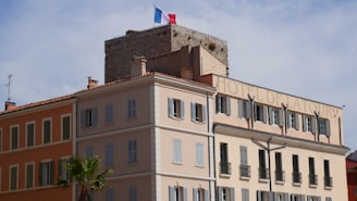 A multi-story building with a light beige and orange facade, featuring numerous windows with closed shutters. The structure has a classic architectural style with a sign reading 'Hotel de la Tour' on the top. A stone watchtower with a French flag on top is visible in the background. Palm trees are seen in the foreground, and the sky is clear with a few clouds.