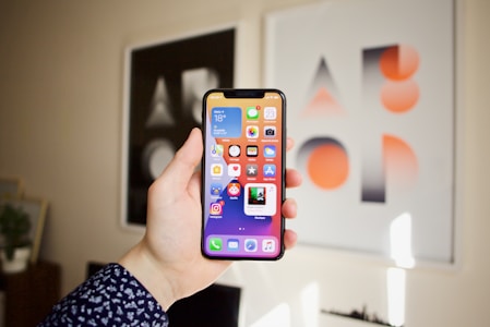 A hand is holding a smartphone displaying various app icons on the home screen. The background features two framed abstract art pieces with geometric designs in black, white, and orange.